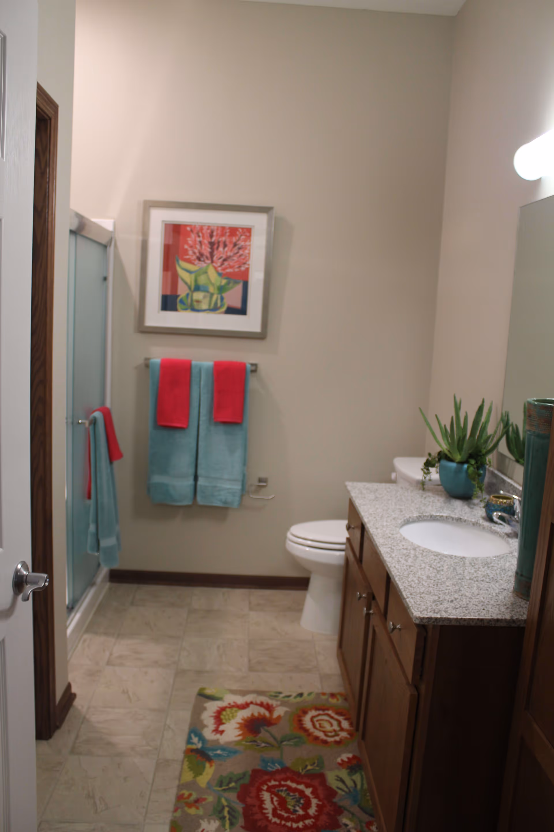 A bathroom with a granite countertop vanity, a white sink, a toilet, and a shower with frosted glass doors. There are two turquoise towels with red hand towels hanging on a towel rack, a colorful floral rug on the tiled floor, a framed artwork of flowers on the wall, and a potted plant on the countertop.