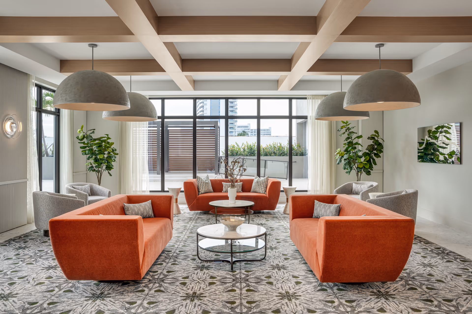A modern living room with three orange sofas arranged around two round marble coffee tables on a patterned carpet. The room features large windows with sheer curtains, two green plants in the corners, and four large dome-shaped pendant lights hanging from a ceiling with wooden beams.