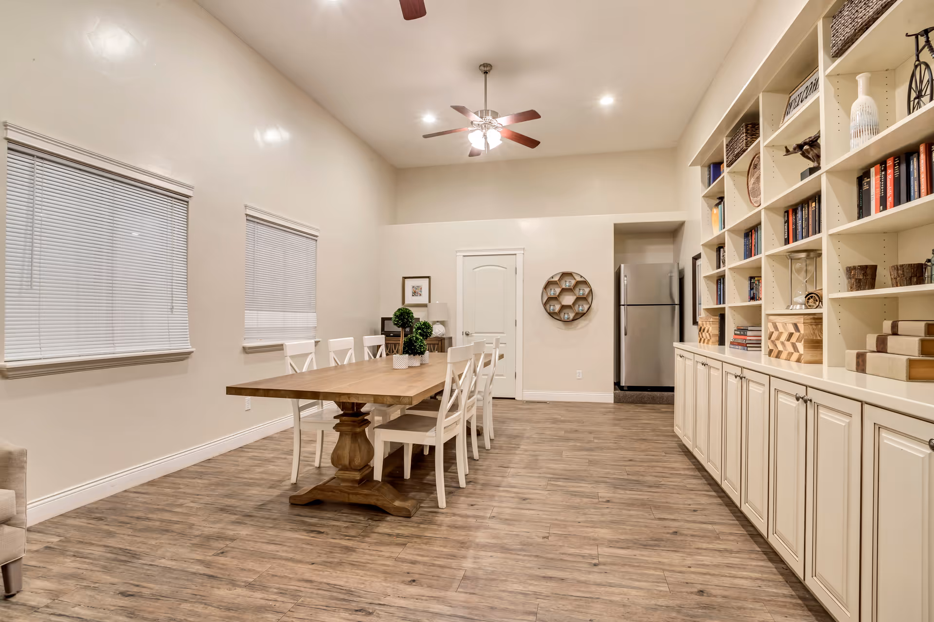 A spacious dining room with a long wooden table surrounded by white chairs. The room features wood flooring, two windows with blinds, a ceiling fan with lights, and built-in white shelves filled with books and decorative items. A stainless steel refrigerator is visible in the background near a closed door.
