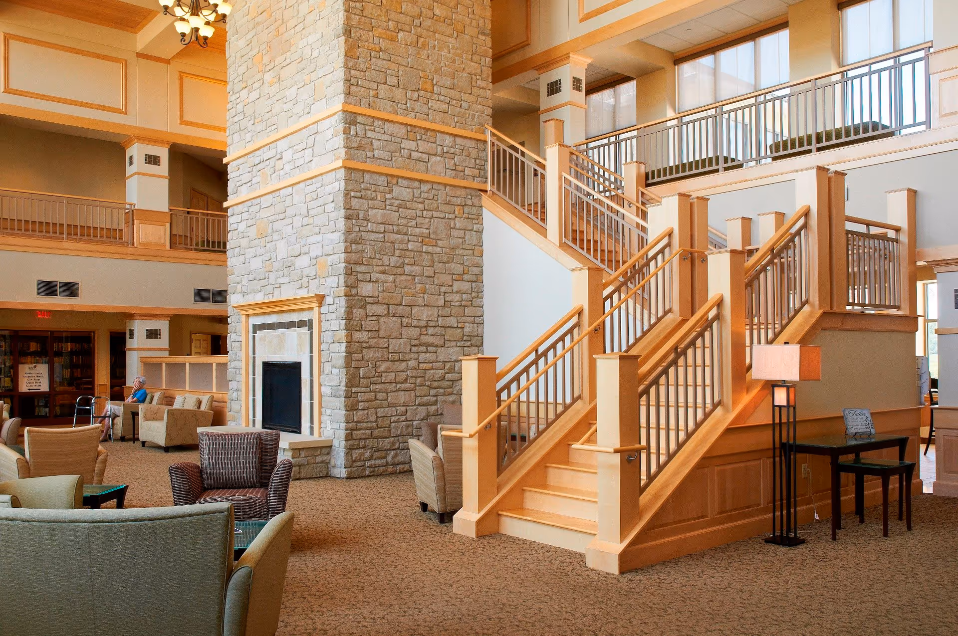 Spacious and well-lit senior living facility common area with a large stone fireplace in the center, surrounded by comfortable seating including armchairs and sofas. A wide staircase with wooden railings leads to an upper level with large windows allowing natural light to fill the space. A floor lamp and small table are positioned near the staircase, and a senior woman is seated in the background near a walker.