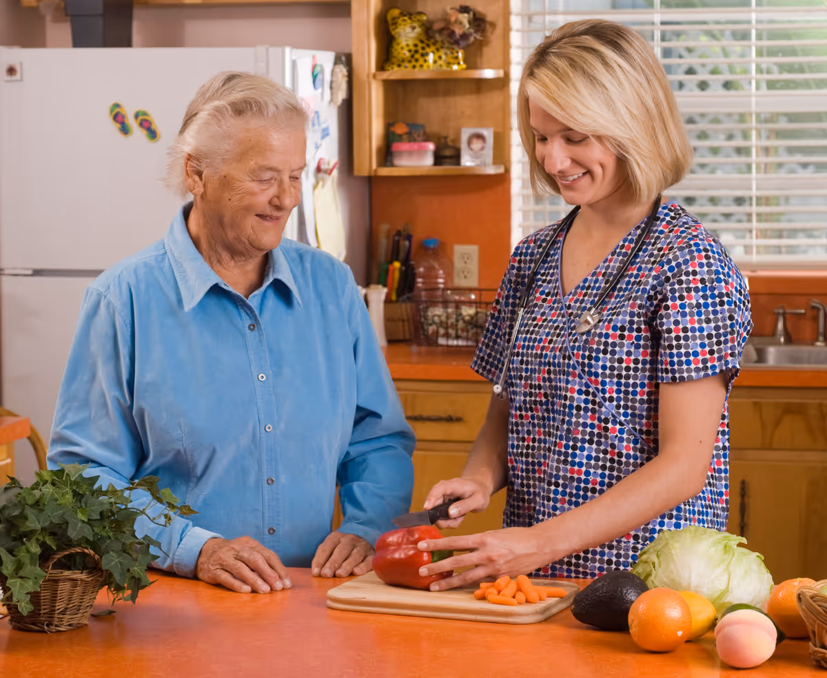A caregiver chops a red bell pepper on a cutting board while an elderly woman watches in a kitchen.