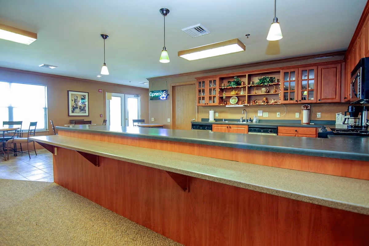 Interior view of a senior living facility kitchen and dining area with a long counter, wooden cabinets, a sink, dishwasher, coffee maker, and tables with chairs near large windows.
