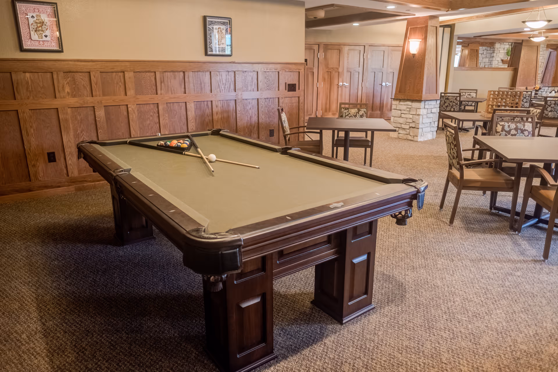 Interior room with a pool table in the foreground, set up with billiard balls and cues. The room has wood paneling on the walls, carpeted floor, and several tables and chairs arranged in the background. The space is warmly lit with wall sconces and ceiling lights.