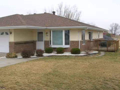 Single-story facility with beige siding, brick accents, front yard, and a wooden deck.