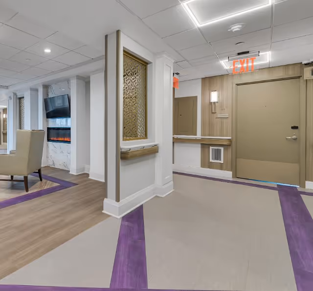 Interior view of a senior living facility hallway with beige walls and ceiling, a beige door with an illuminated exit sign above it, a decorative wooden panel with a geometric pattern, a wall-mounted light fixture, and a seating area with a beige armchair and a wall-mounted TV above an electric fireplace. The floor has a beige and purple patterned carpet.