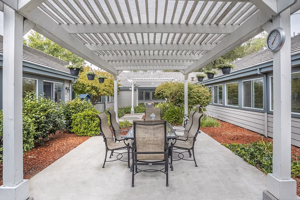 Outdoor patio area with a white pergola overhead, a glass-top table surrounded by six cushioned chairs, and greenery including bushes and hanging plants around the concrete floor. The patio is situated between two buildings with multiple windows.