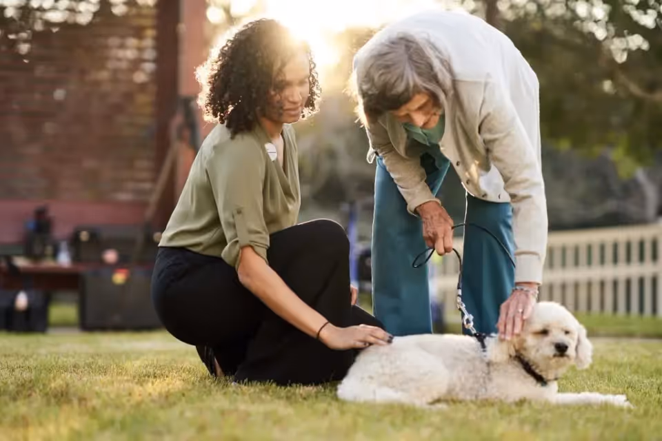 An elderly woman and a younger woman outdoors on grass, interacting with a white dog lying on the ground. The elderly woman is bending over petting the dog while holding its leash, and the younger woman is kneeling nearby, also touching the dog. The background shows a fence and trees with sunlight filtering through.