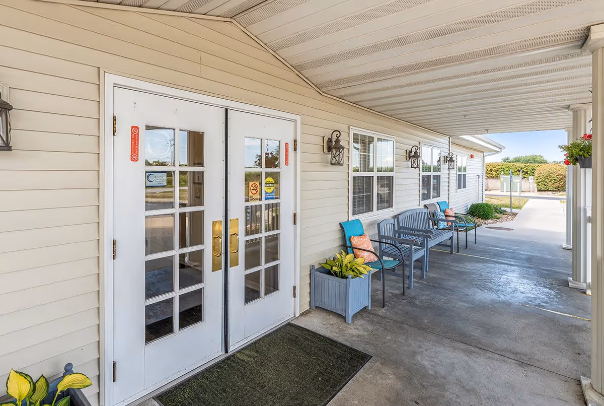 Covered entrance area of a senior living facility with double white doors featuring glass panes and brass handles. Along the wall, there are several gray benches with blue cushions and orange patterned pillows, accompanied by a planter with green and yellow foliage. The area is well-lit with wall-mounted lantern-style lights and has a concrete floor extending to a driveway or parking area.