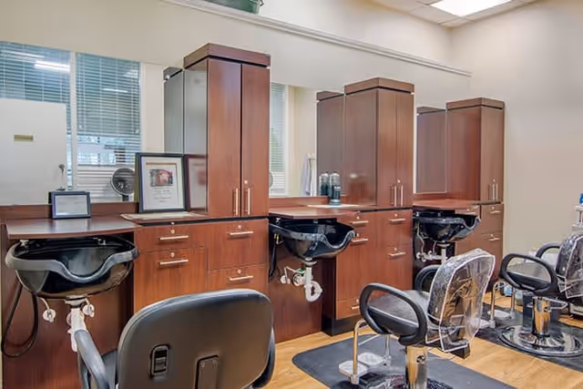 Interior view of a salon area with three black salon chairs in front of wooden cabinetry and sinks for hair washing. The cabinetry has multiple drawers and cabinets, and there are mirrors above the sinks. The floor is wooden, and the room is well-lit with windows in the background.