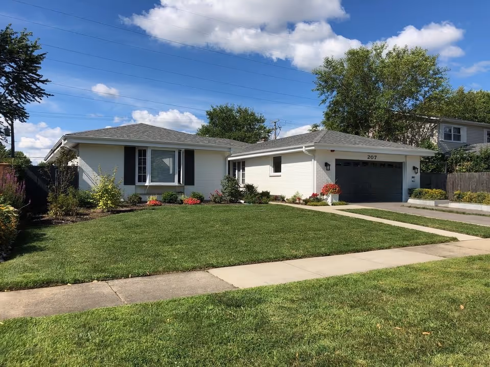 Single-story white brick house with a gray shingled roof, a large front lawn, and a driveway leading to a dark gray garage door. The house has a large window with black shutters and is surrounded by small bushes and flowers. The sky is blue with some clouds.