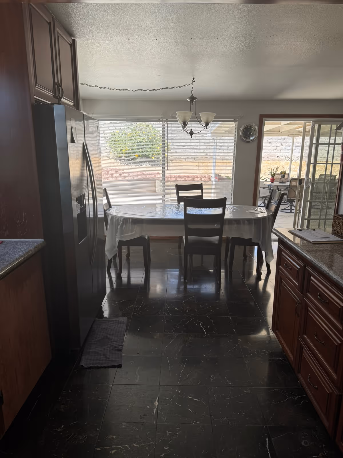 Dining area with a rectangular table covered with a white tablecloth and four wooden chairs. The room has large windows with blinds letting in natural light, a hanging light fixture above the table, and a sliding glass door leading to an outdoor patio. The floor is dark marble tile, and there are wooden cabinets and a stainless steel refrigerator visible on the sides.