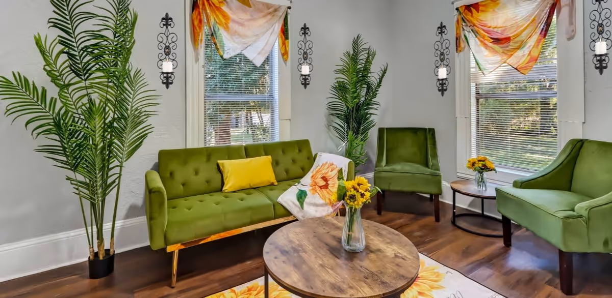 A cozy living room with green velvet sofa and chairs, a wooden round coffee table with a vase of yellow flowers, two large windows with floral curtains, decorative wall sconces with candles, and two tall potted plants in the corners.