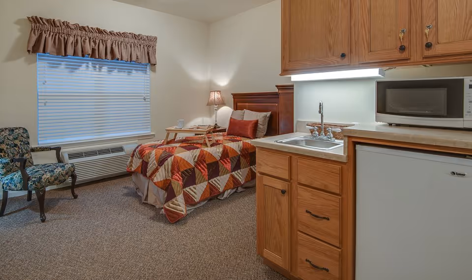 A cozy senior living room with a single bed covered in a colorful patchwork quilt, a wooden headboard, and a bedside table with a lamp and a tray. To the right, there is a kitchenette area with wooden cabinets, a small sink, a microwave, and a mini refrigerator. A floral upholstered armchair is placed near a window with closed blinds and a brown valance.