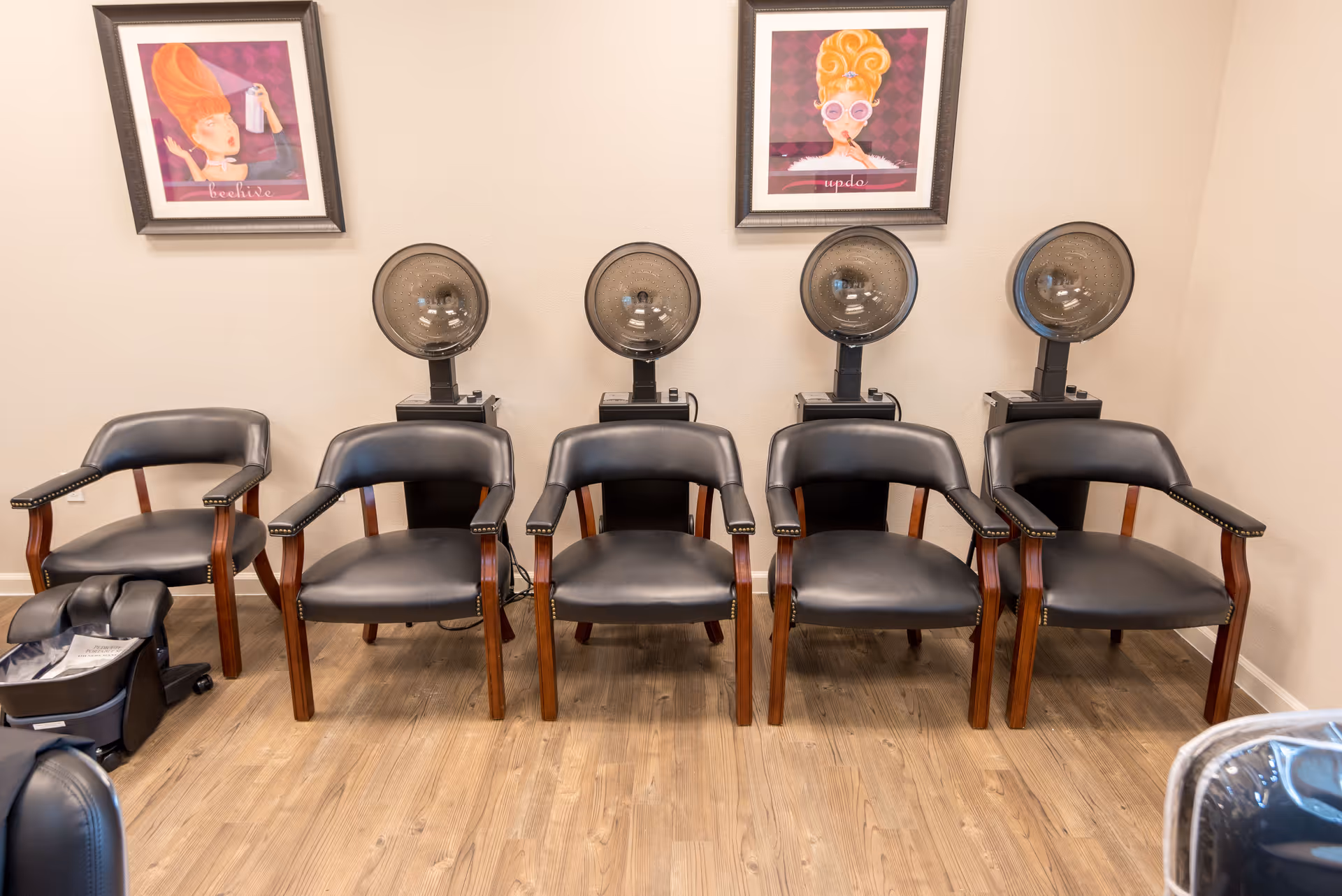 A row of five black salon chairs with wooden armrests positioned in front of four vintage hair dryers in a salon or beauty area. Two framed colorful illustrations of women with styled hair hang on the beige wall above the hair dryers. The floor is wooden.