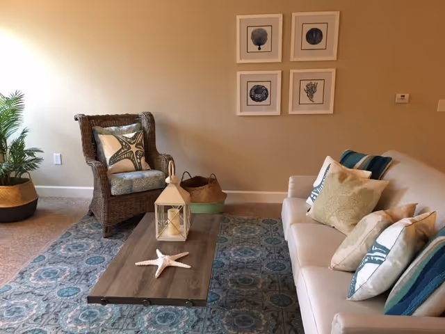 A cozy living room with a white sofa adorned with multiple decorative pillows, a wicker armchair with a starfish-patterned pillow, a wooden coffee table with a starfish decoration and a lantern, a patterned blue area rug, a potted plant in the corner, and four framed sea-themed artworks on the beige wall.
