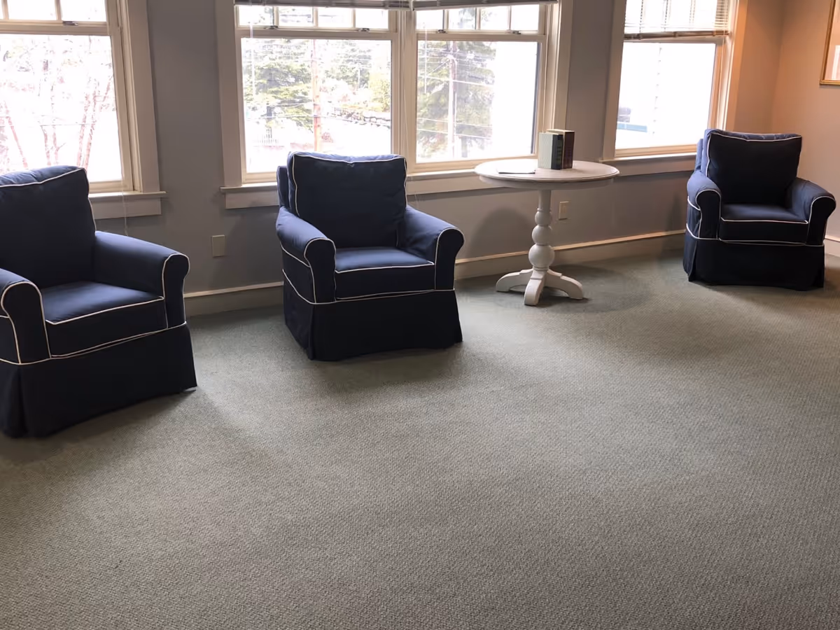 A room with three navy blue armchairs with white piping arranged in a row in front of three large windows. There is a small round white table between the middle and right armchairs with a few books on top. The room has light-colored walls and a light green carpeted floor.