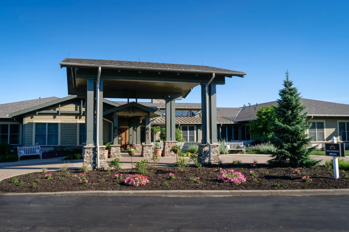 Front entrance of a single-story senior living building with a covered porte-cochere, landscaped flower beds, benches, and a main entrance sign.