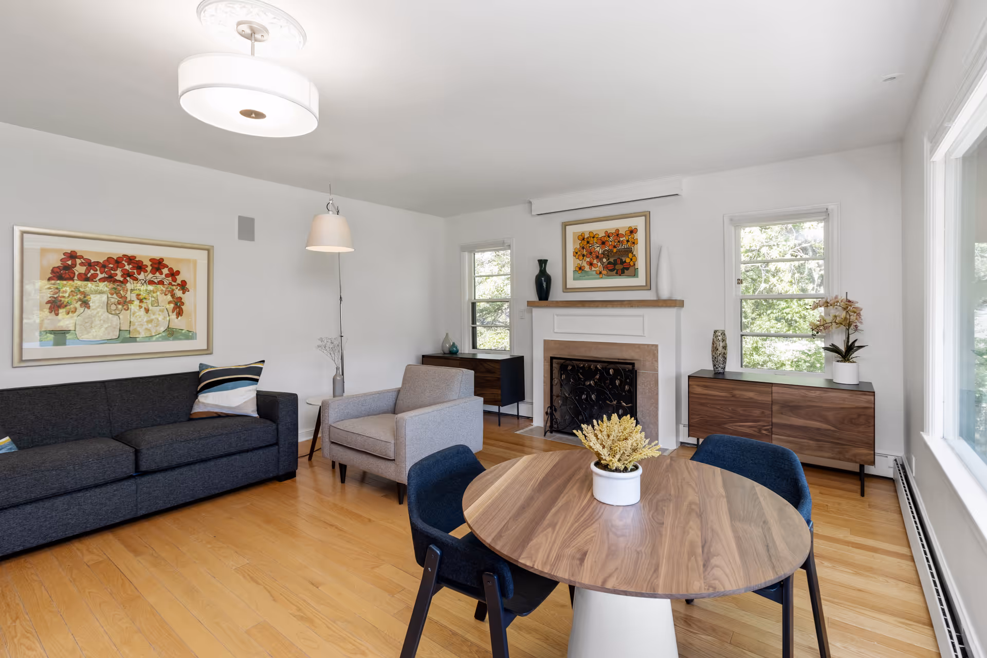 A bright living room with light wood flooring, a dark gray sofa, a gray armchair, a round wooden table with four dark chairs, and a white planter with yellow flowers on the table. The room features a white fireplace with decorative vases and framed floral artwork above it. Two windows flank the fireplace, and there is a large window on the right side letting in natural light. The walls are white, and there is a ceiling light fixture and a hanging lamp near the armchair.