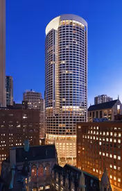 Tall cylindrical skyscraper building illuminated at dusk, surrounded by other city buildings under a clear blue sky.