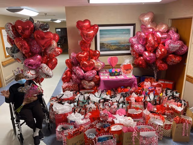 A hallway in a senior living facility decorated with numerous Valentine's Day themed gift bags and heart-shaped balloons in red, pink, and white colors. A person in a wheelchair is partially visible on the left side holding some balloons. A table covered with a pink cloth holds additional Valentine's Day decorations and cards. A framed landscape painting hangs on the wall behind the table.