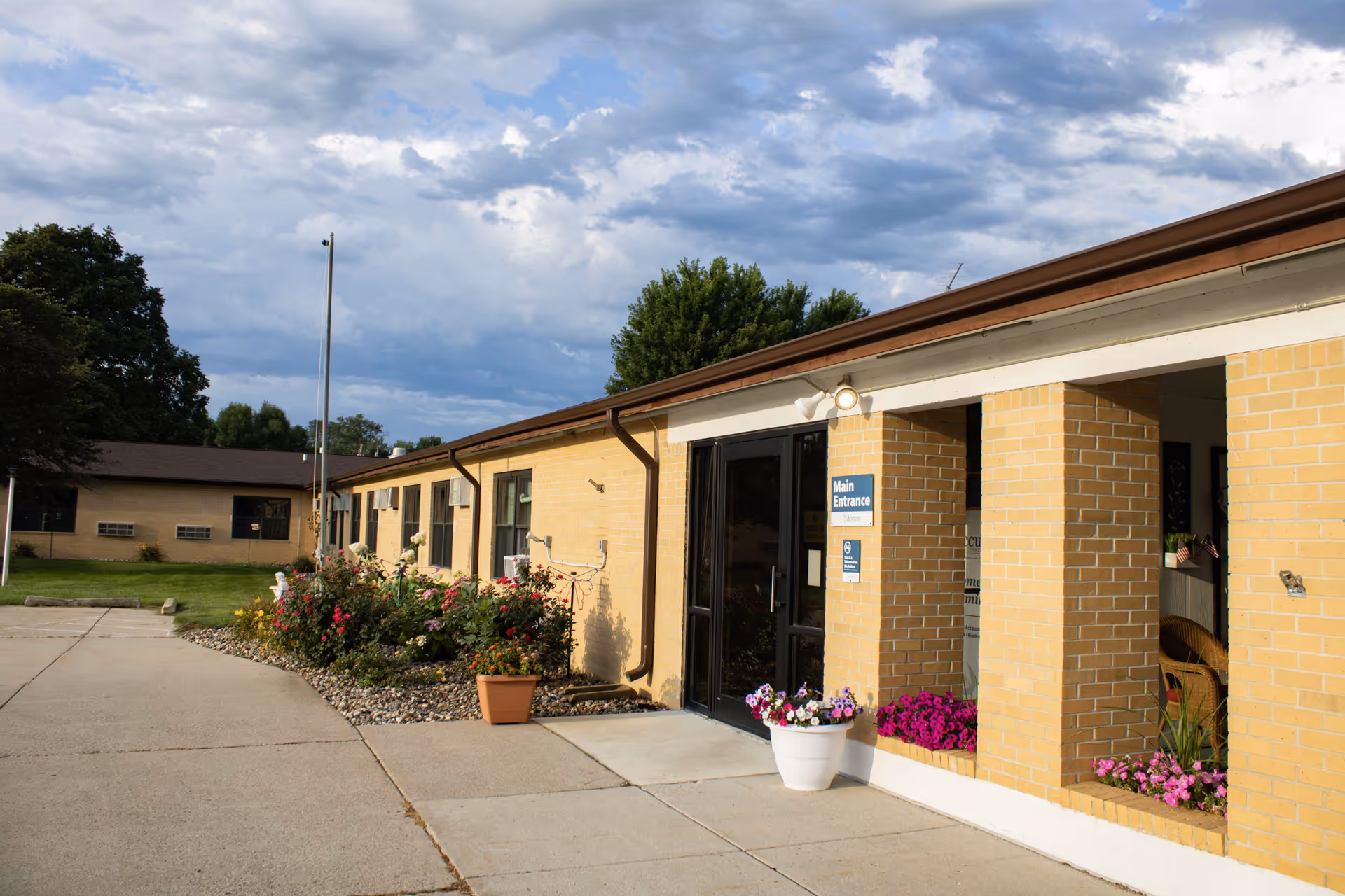 Front entrance of a single-story yellow brick building with potted flowers and a walkway under a cloudy sky.