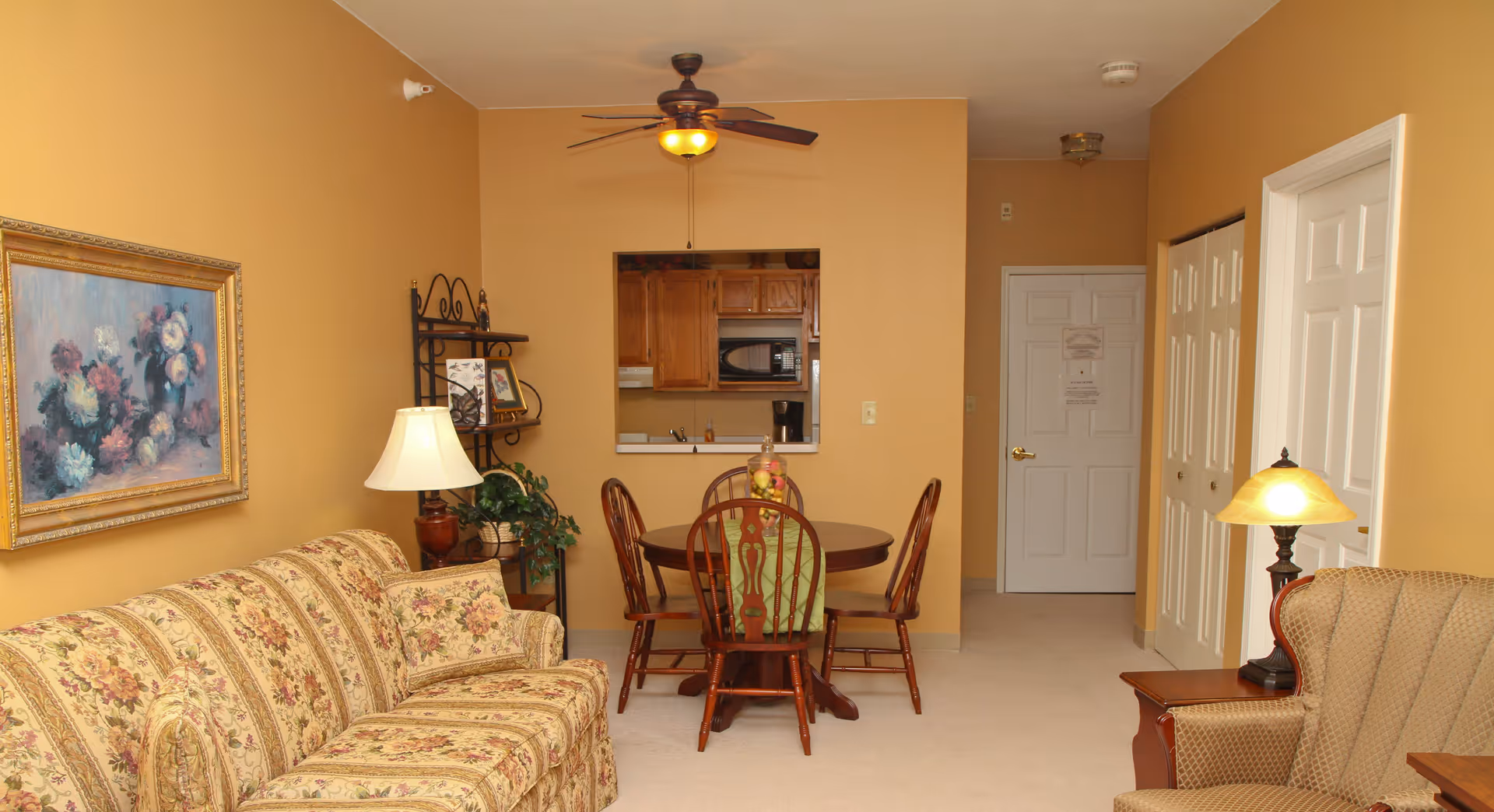 A cozy living and dining area with mustard yellow walls. The living room features a floral patterned sofa, a side table with a lamp, and an armchair with another lamp beside it. A framed floral painting hangs on the wall above the sofa. The dining area has a round wooden table with four chairs and a fruit centerpiece. A ceiling fan with a light is mounted above the dining table. In the background, there is a small kitchen area visible through a pass-through window, with wooden cabinets and a microwave. The hallway leads to a white door and closets with white folding doors.