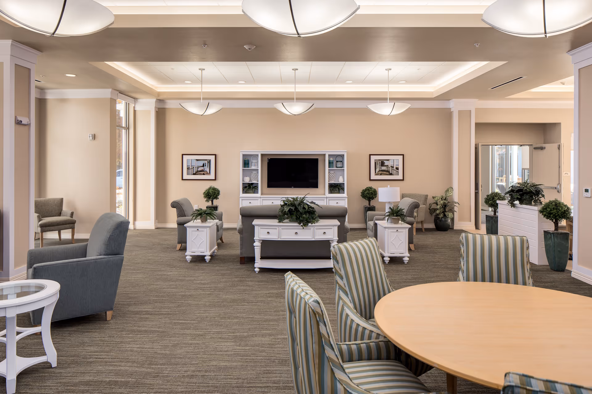 A spacious and well-lit living room area in a senior living facility with beige walls and carpeted floors. The room features multiple comfortable armchairs and sofas arranged around white wooden tables with green plants on top. A flat-screen TV is mounted on the far wall, flanked by shelves with decorative items. There are framed pictures on the walls and large windows letting in natural light. A round wooden table with striped upholstered chairs is in the foreground.