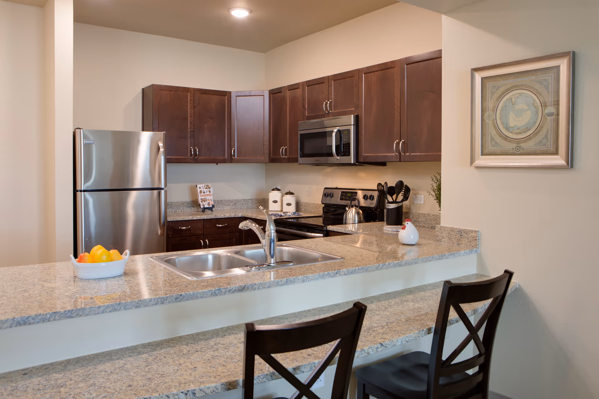 Modern kitchen with granite countertops, a double sink, stainless steel refrigerator, microwave, and stove. Dark wood cabinets line the walls, and two dark wood chairs are positioned at the counter. A framed artwork hangs on the wall to the right.