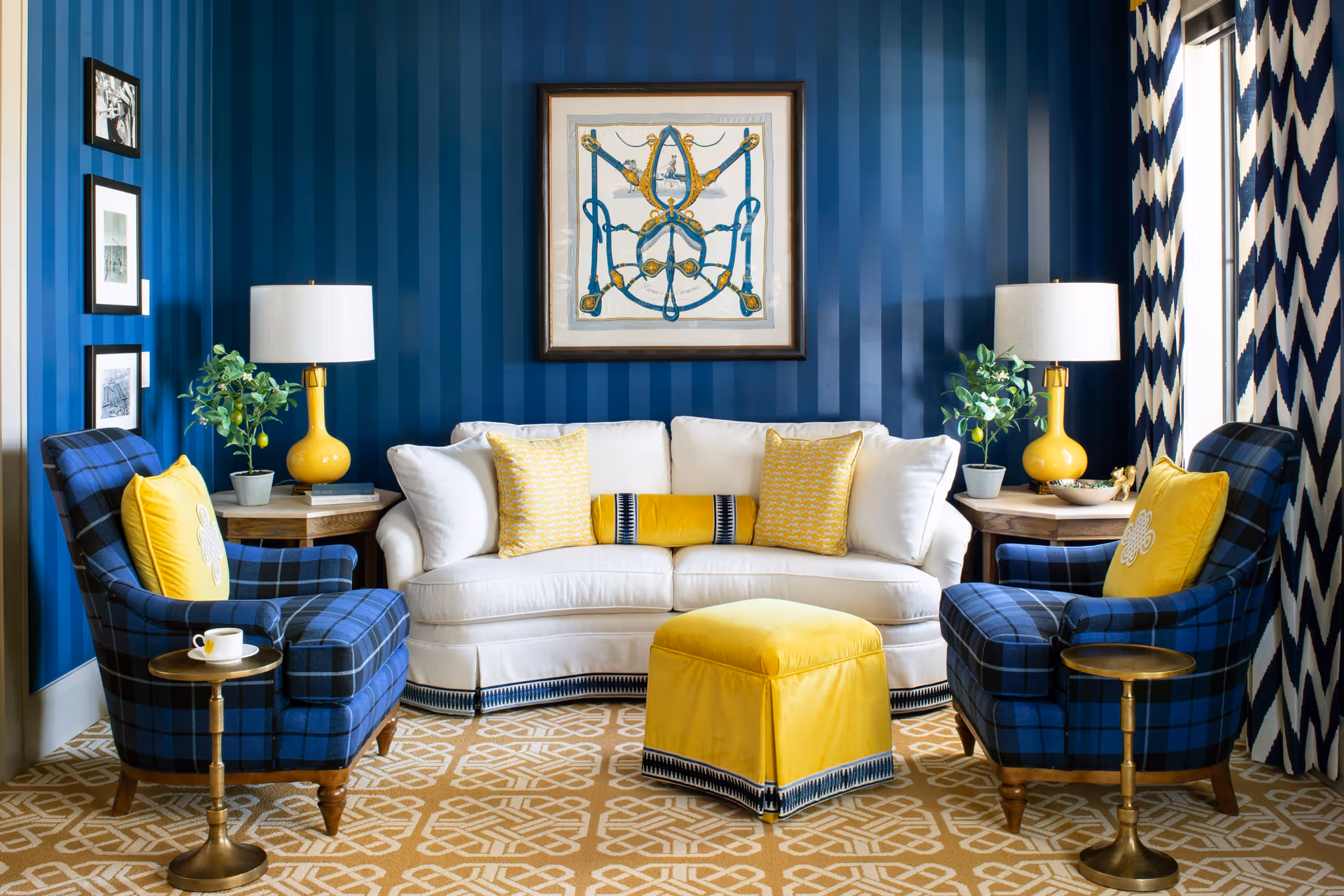 A cozy living room with a white sofa adorned with yellow and white pillows, flanked by two blue plaid armchairs each with a yellow pillow. Between the armchairs and sofa are wooden side tables with yellow lamps and small green plants. A yellow ottoman sits in front of the sofa on a beige patterned carpet. The walls are painted in dark blue vertical stripes, and a framed artwork hangs above the sofa. Blue and white chevron curtains cover the window on the right side.
