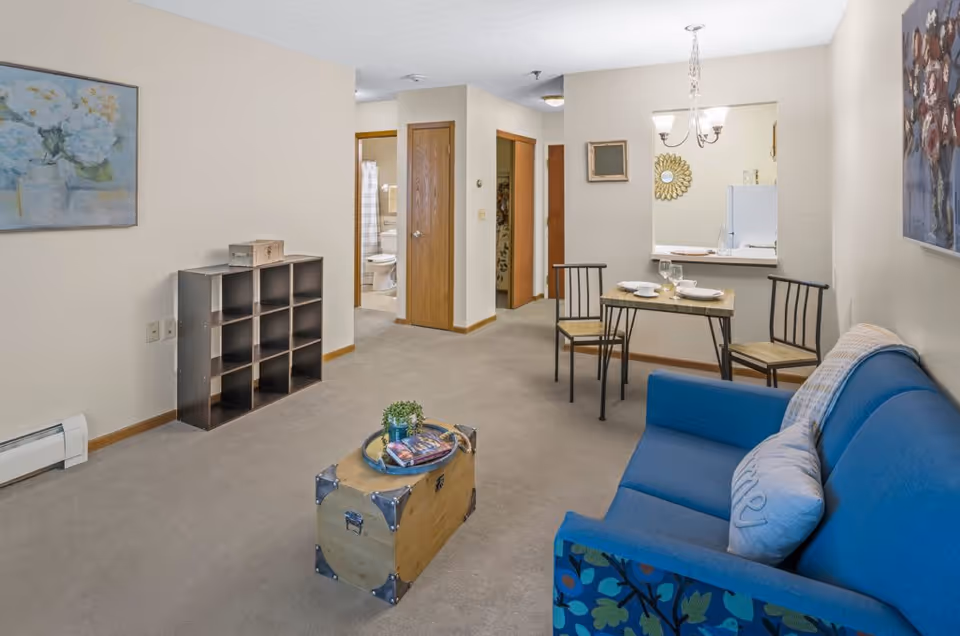 A cozy living room area with a blue couch adorned with pillows, a small wooden chest used as a coffee table with a plant and magazines on it, and a black cubby shelf against the wall. In the background, there is a small dining table set for two with plates and glasses, and an open kitchen pass-through window. The walls are decorated with framed artwork, and a bathroom is visible through an open door.