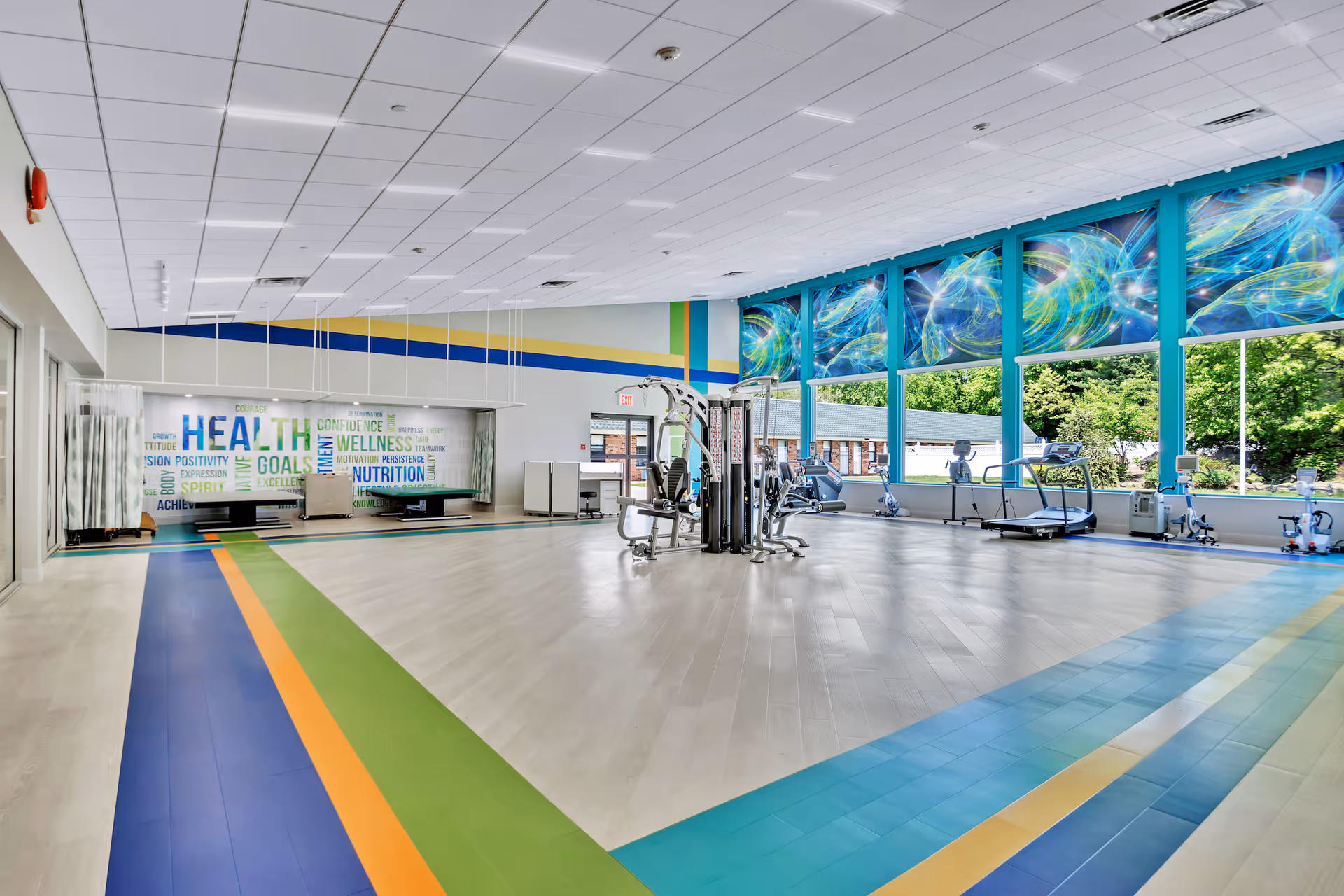 A spacious and brightly lit fitness room with exercise equipment including treadmills and weight machines. The room features large windows with colorful abstract designs at the top, allowing natural light to enter and offering a view of greenery outside. The floor has colorful stripes in blue, green, and orange, and a wall with motivational words related to health and wellness.