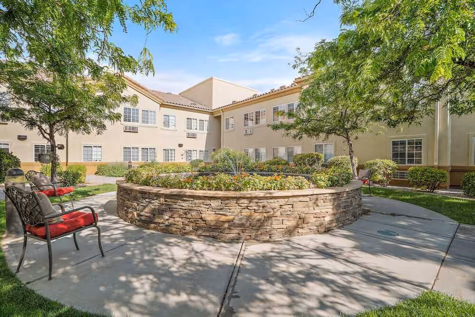 Sunny courtyard with a circular raised stone planter, trees, and metal benches with red cushions in front of a two-story beige building.