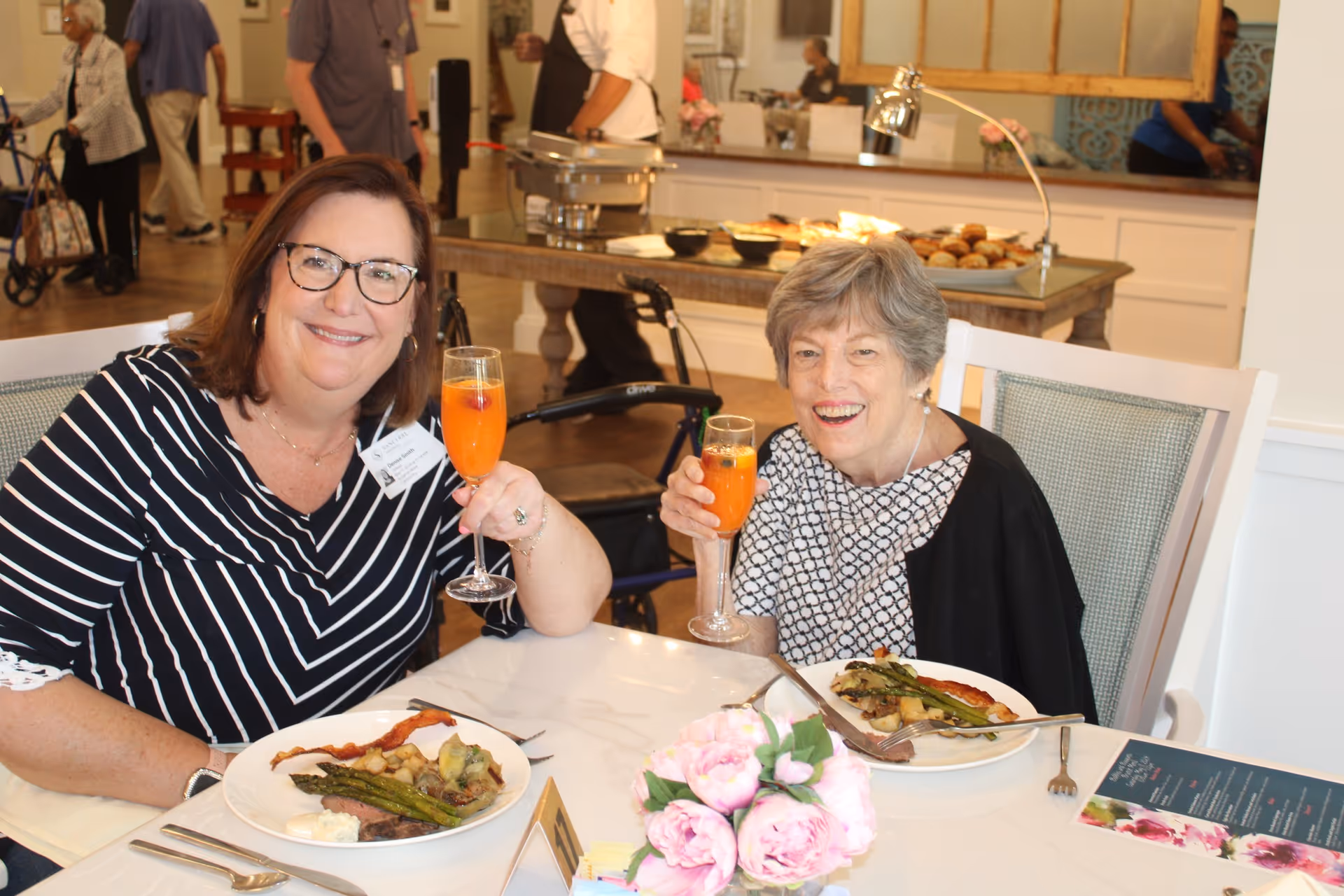 Two women sitting at a dining table in a senior living facility, smiling and holding glasses of orange-colored drinks. Plates with food including asparagus, bacon, and potatoes are in front of them. The background shows other people and a buffet table with food.