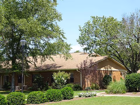 Single-story brick building with a tan roof surrounded by green trees, bushes, and a well-maintained lawn under a clear blue sky.