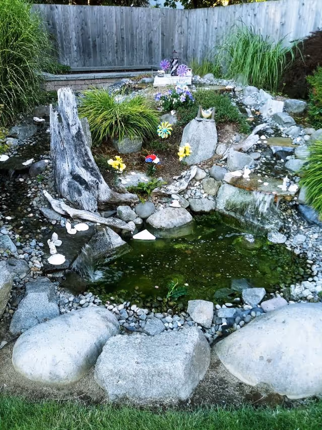 A small landscaped garden pond surrounded by rocks and greenery, featuring a wooden log, decorative flowers, small figurines, and a wooden fence in the background.