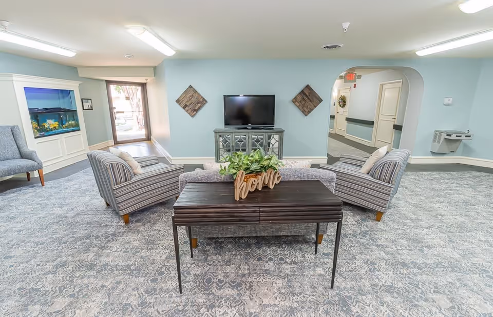A senior living facility common area with light blue walls and patterned carpet. The room features a flat-screen TV on a cabinet against the wall, two striped armchairs, and a sofa arranged around a dark wooden table with a decorative 'home' sign and a plant. There is an aquarium built into the wall on the left side and a water fountain on the right. A doorway and hallway are visible in the background.