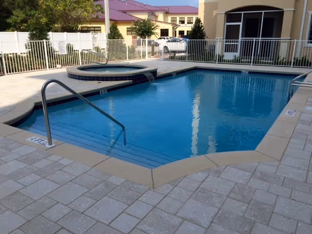 Outdoor swimming pool with clear blue water and a connected circular hot tub, surrounded by a tiled deck and white safety fence. In the background, there are buildings with beige walls and purple roofs, along with some trees and parked cars.