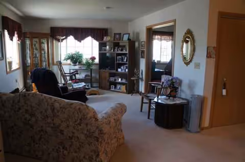 Cozy living room with floral sofa, chairs, bookshelf, plants, and large windows.