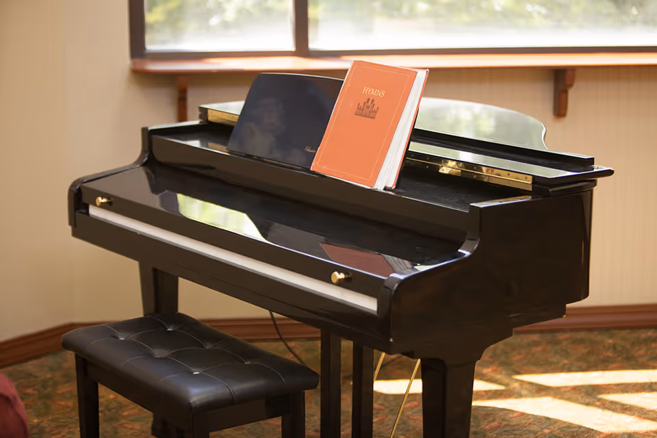 Black grand piano with a padded bench and an open hymns book on the music rack in a sunlit room.