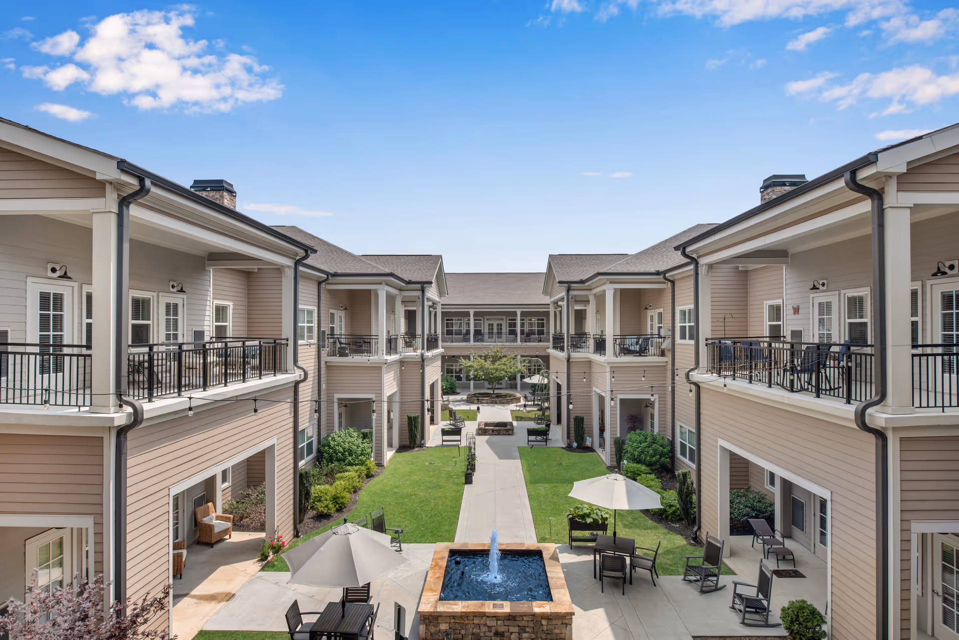 Courtyard of a senior living facility with a central fountain, patio seating and umbrellas surrounded by two-story buildings with balconies.