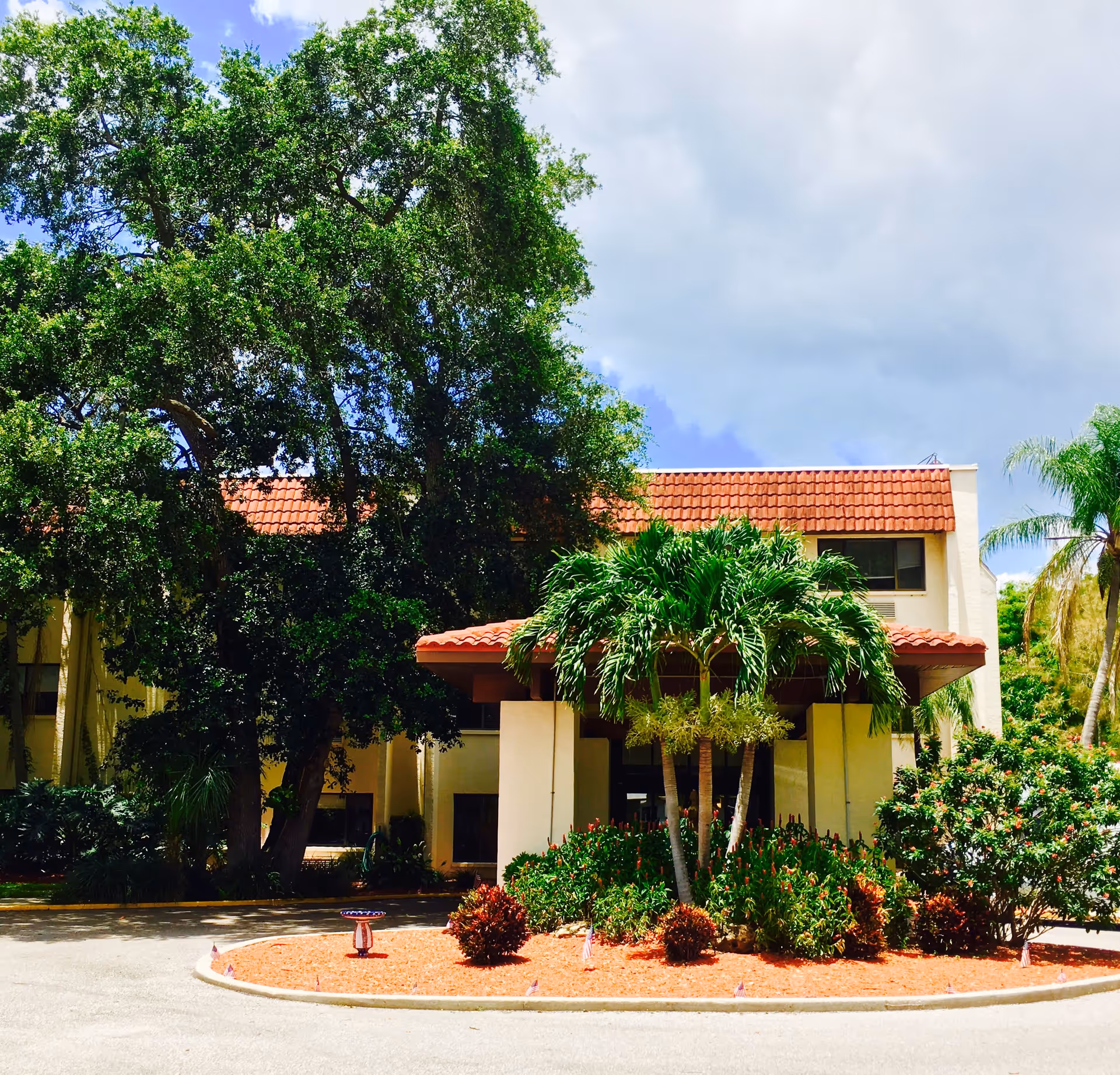 Front entrance of a two-story building with a red-tiled roof, palm trees, and a landscaped circular driveway.
