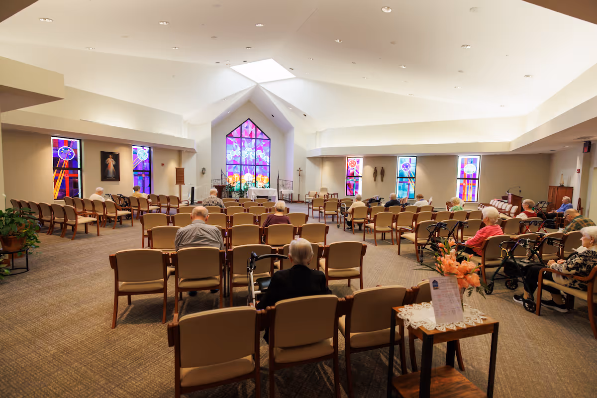 Interior view of a chapel or worship room in a senior living facility with rows of chairs arranged facing a stained glass window at the front. Several elderly individuals are seated, some using walkers. The room is well-lit with a high ceiling and additional stained glass windows on the side walls.