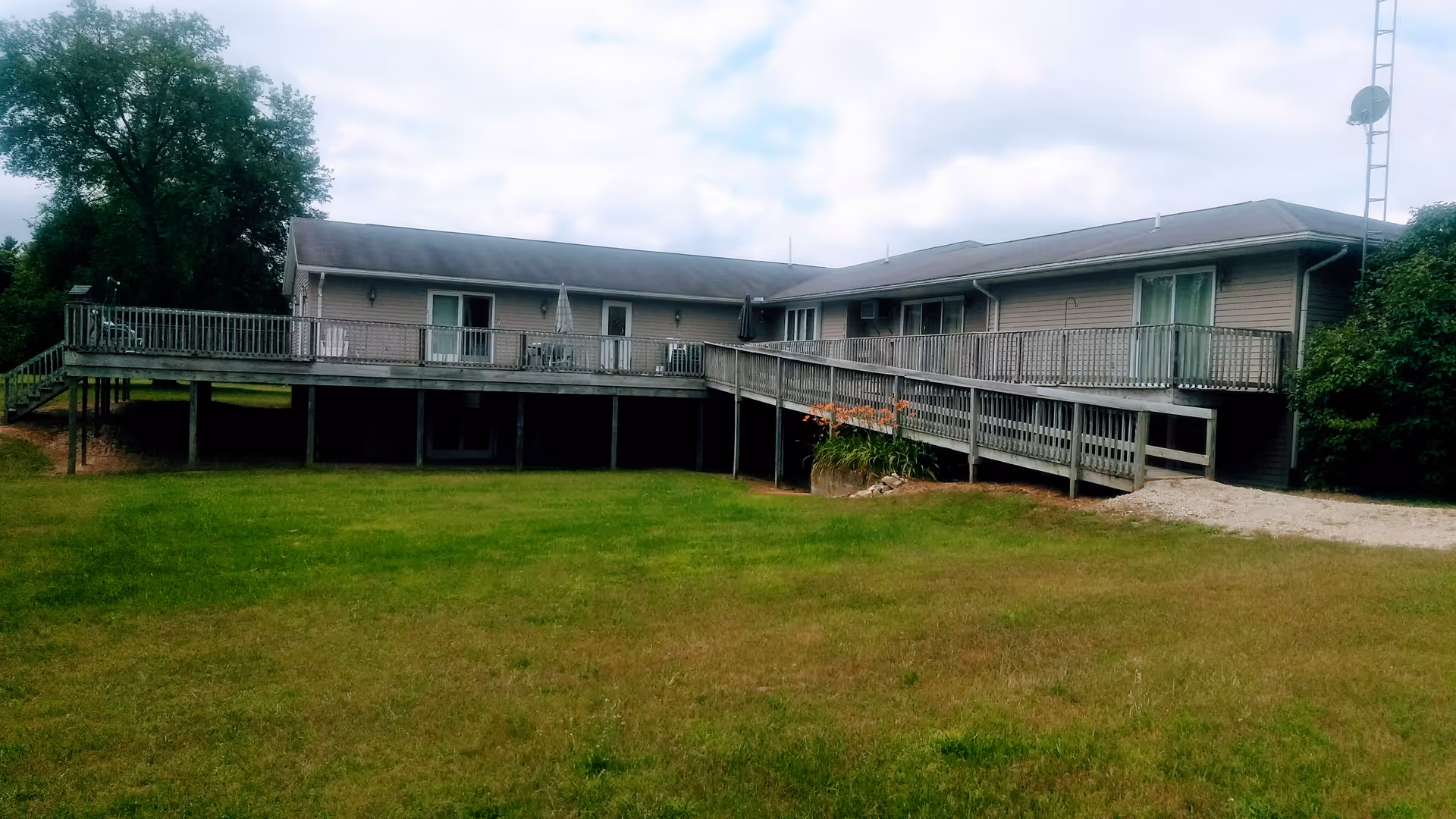Exterior view of a single-story building with a large wooden deck and wheelchair ramp, surrounded by a grassy lawn and trees under a cloudy sky.