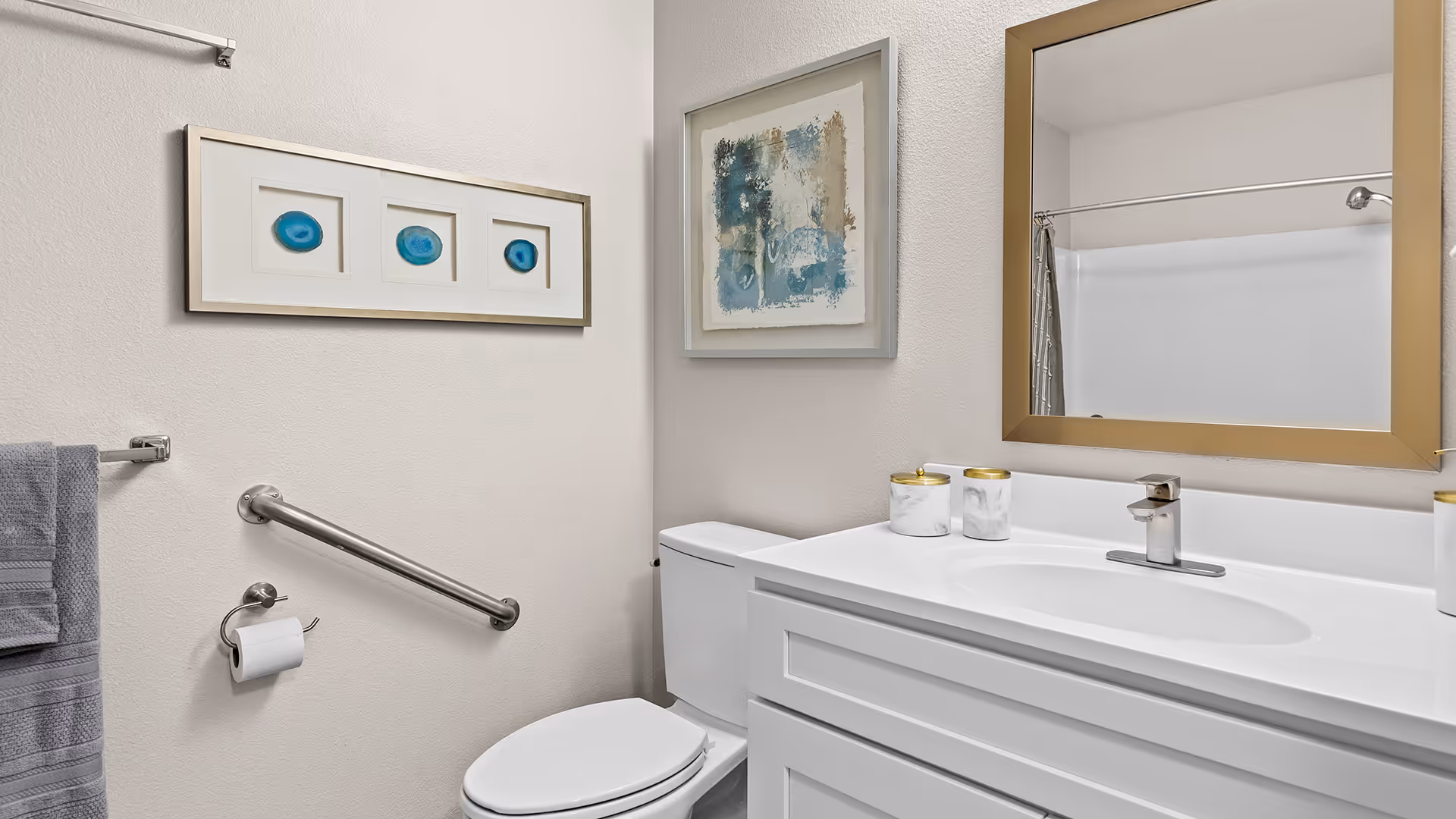 A clean and modern bathroom featuring a white toilet, white vanity with a sink, and a large mirror with a gold frame. The wall has two pieces of framed artwork, one with blue circular designs and the other with abstract blue and beige patterns. There is a metal grab bar next to the toilet, a towel rack with a gray towel, and a toilet paper holder. A shower curtain and showerhead are partially visible in the mirror reflection.