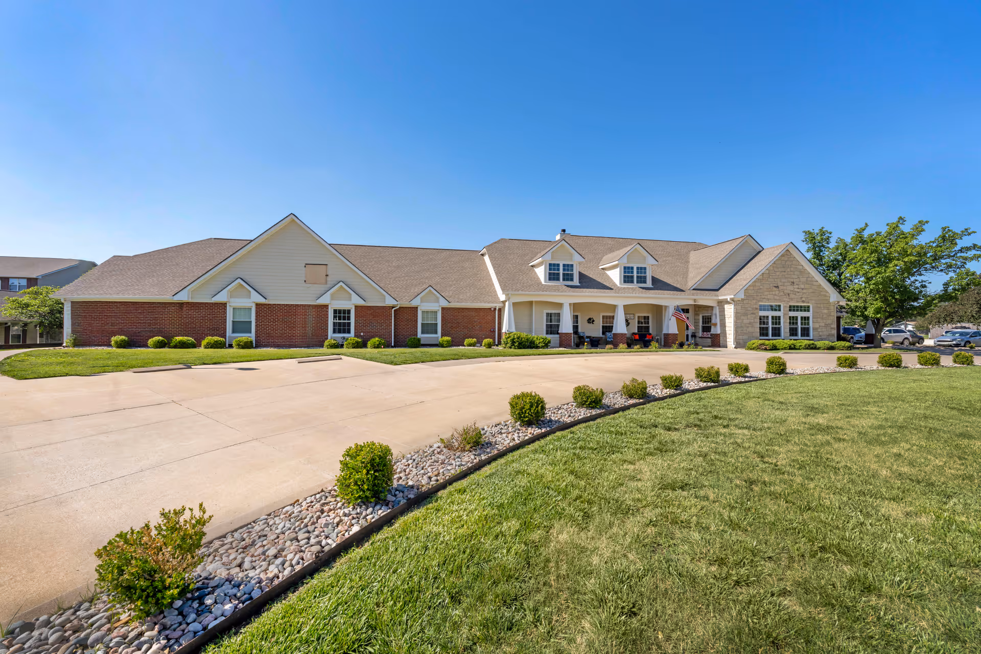 Exterior view of a single-story senior living facility building with a combination of brick and stone facade, a covered entrance with white columns, an American flag, and a well-maintained lawn with small bushes and a rock border under a clear blue sky.
