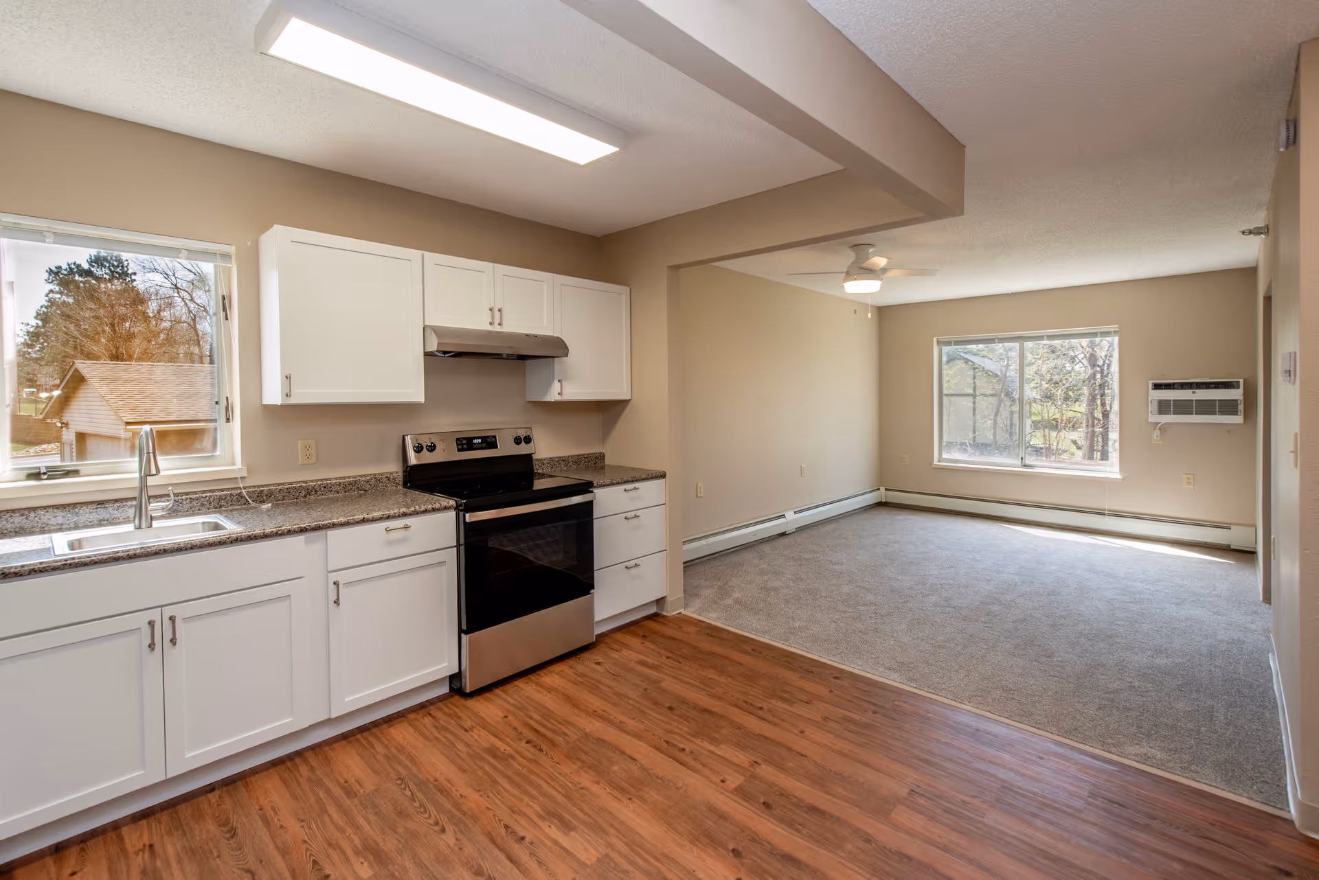 Empty open-concept apartment kitchen with white cabinets, stainless steel stove, wood floors and an adjoining carpeted living area.