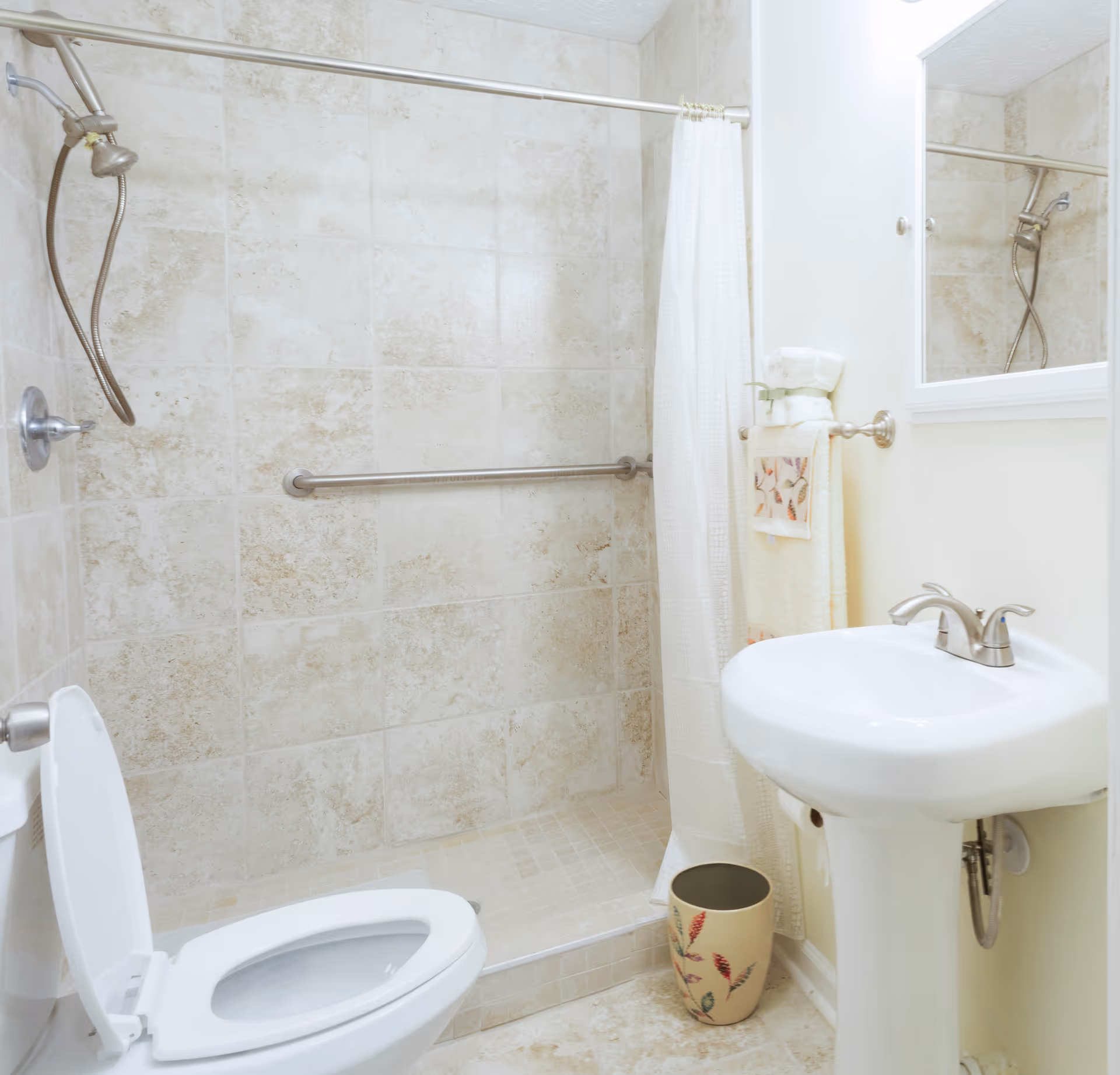 A clean bathroom with a tiled walk-in shower featuring a handheld showerhead and a grab bar. There is a white toilet with the lid open, a white pedestal sink with a silver faucet, a mirror above the sink, and a towel rack holding folded towels and a decorative hand towel. A small decorative waste bin is placed on the floor near the sink.