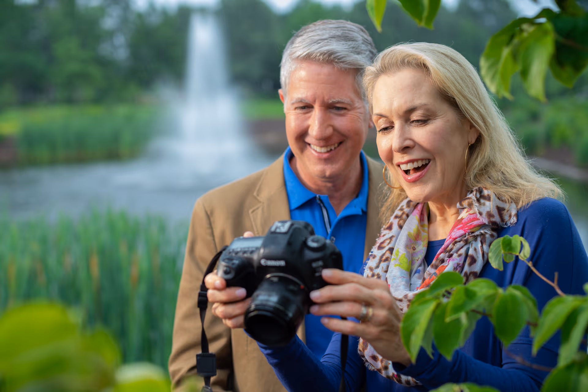 A smiling older couple outdoors near a pond with a fountain, looking at a camera together surrounded by greenery.