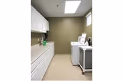 A laundry room with white cabinets and countertop on the left side, and a washing machine and dryer on the right side. The walls are painted olive green, and there is a window and a ceiling light providing illumination.