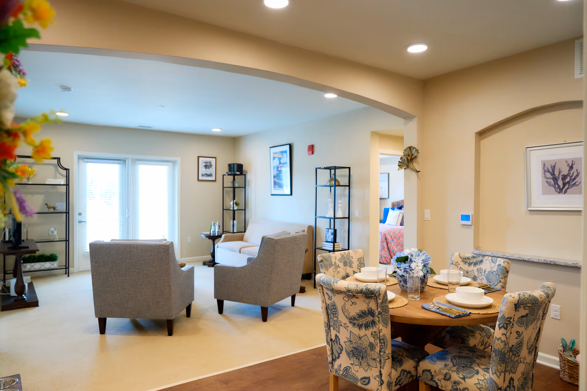 A bright and cozy living and dining area in a senior living facility. The living room features two gray armchairs and a beige loveseat arranged around a small round table. The dining area has a round wooden table set with white dishes and floral-patterned upholstered chairs. The walls are painted a warm beige, and there are framed pictures and decorative shelves. French doors with blinds let in natural light.